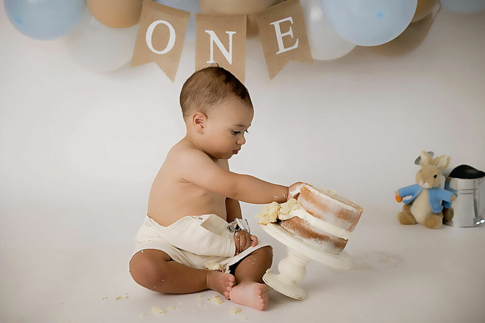 One year old boy, sitting on floor and tipping over for his birthday cake for his first birthday photo shoot taken in Moorestown, New Jersey.