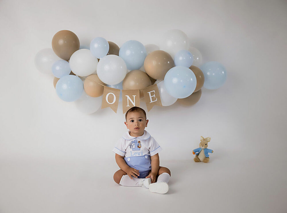 One year old boy, sitting down, looking at camera with balloon, decorations and banner that says one next to Peter rabbit plushy for his Peter rabbit, first birthday session, taken in Southampton, New Jersey.