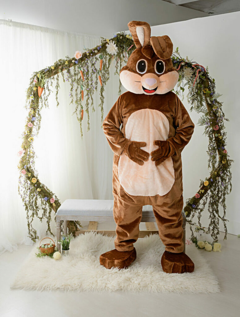 Man in Easter bunny suit posing for the camera while holding belly standing in a photography studio in front of Easter photography prop for mini session promotion in Cherry Hill, New Jersey.