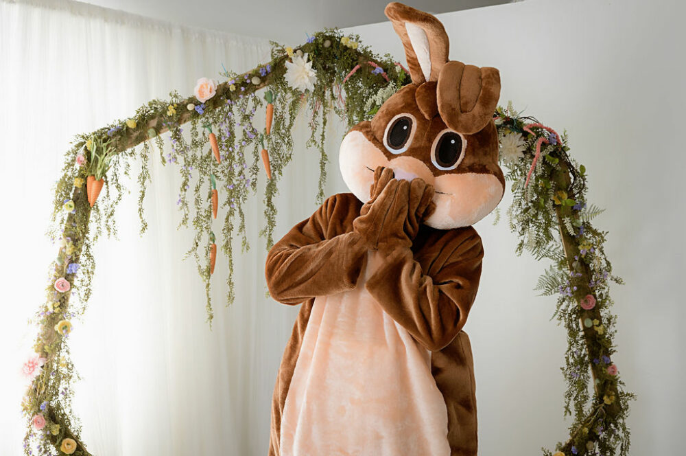 Brown Easter bunny posing for camera covering mouth with hands standing in front of Easter photography prop in Lumberton, New Jersey.