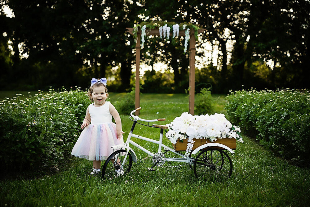 Toddler girl, smiling, a camera standing next to bike, photography prop, standing outdoors and flower garden, port, her outdoor second birthday session in Southampton, New Jersey.