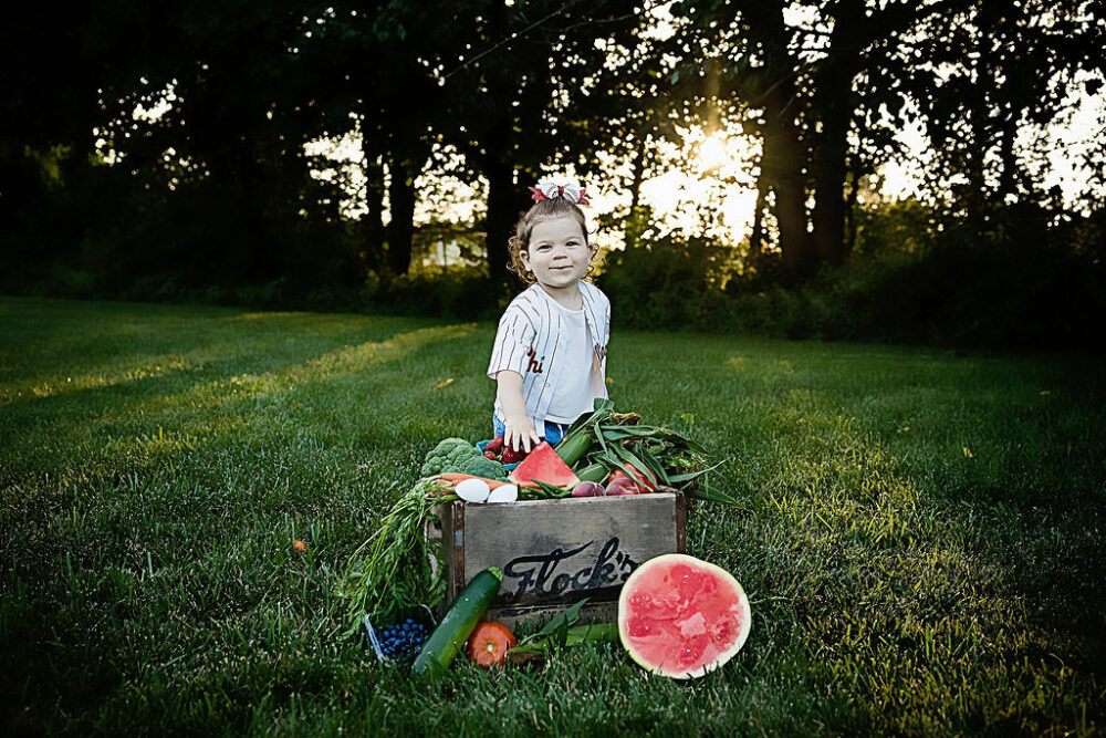 Two-year-old girl posing for camera behind veggie photography props for her best baby photos taken in Eastampton, New Jersey.