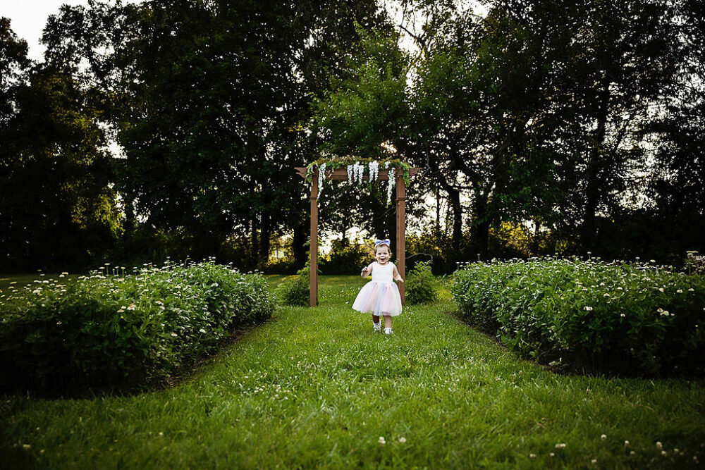 Young girl, smiling and running toward camera and flower garden for her outdoor second birthday session, taken in Camden, New Jersey.