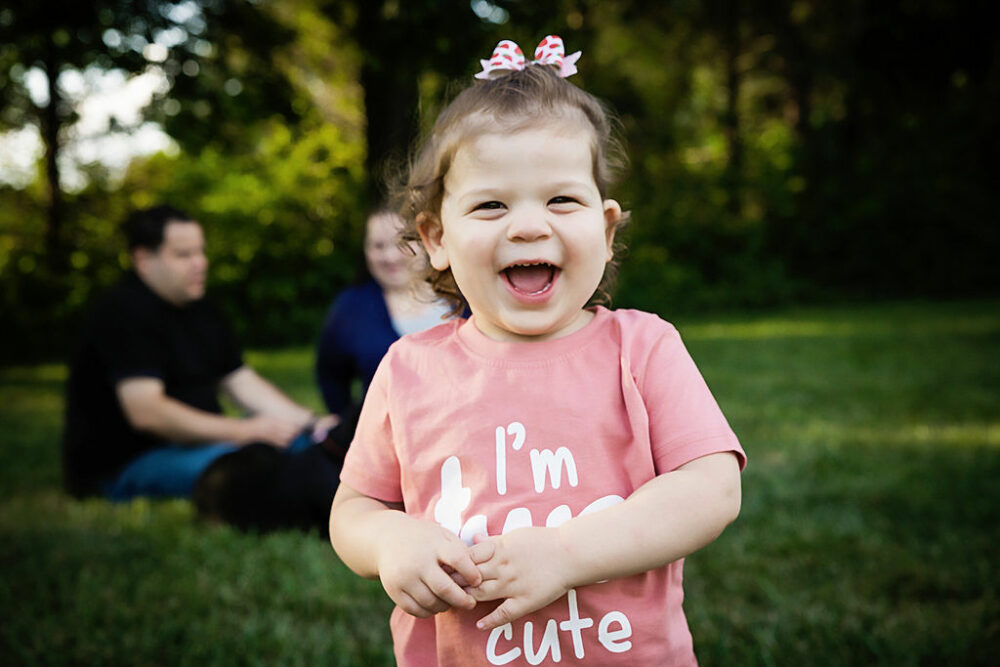 Toddler girl, looking at camera and smiling with her parents in the background sitting on the grass for her summer outdoor second birthday session, taken in Haddonfield, New Jersey.