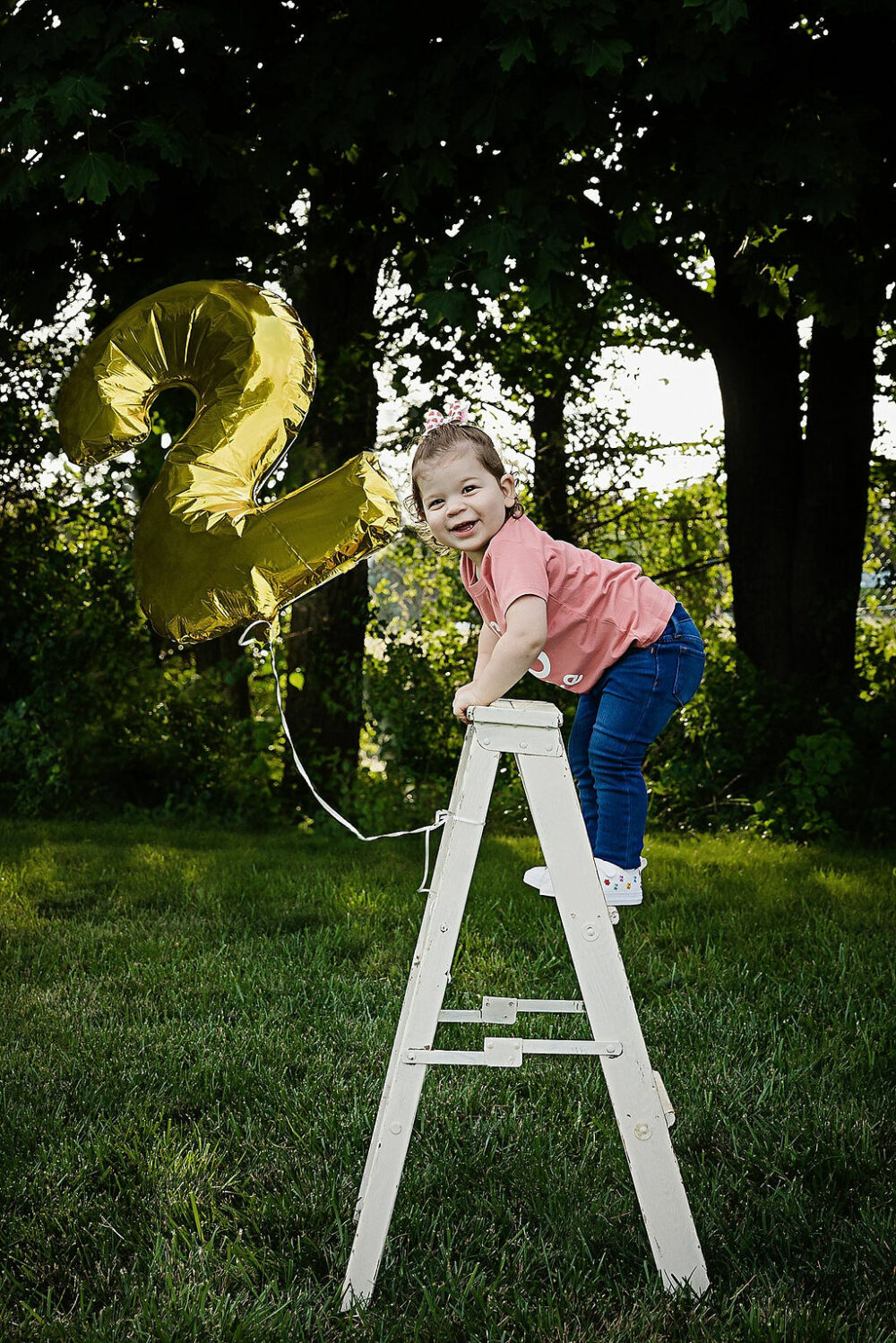 Young girl standing on ladder prop with a balloon number two attached, smiling at camera for her outdoor second birthday session, taken in freehold, New Jersey.