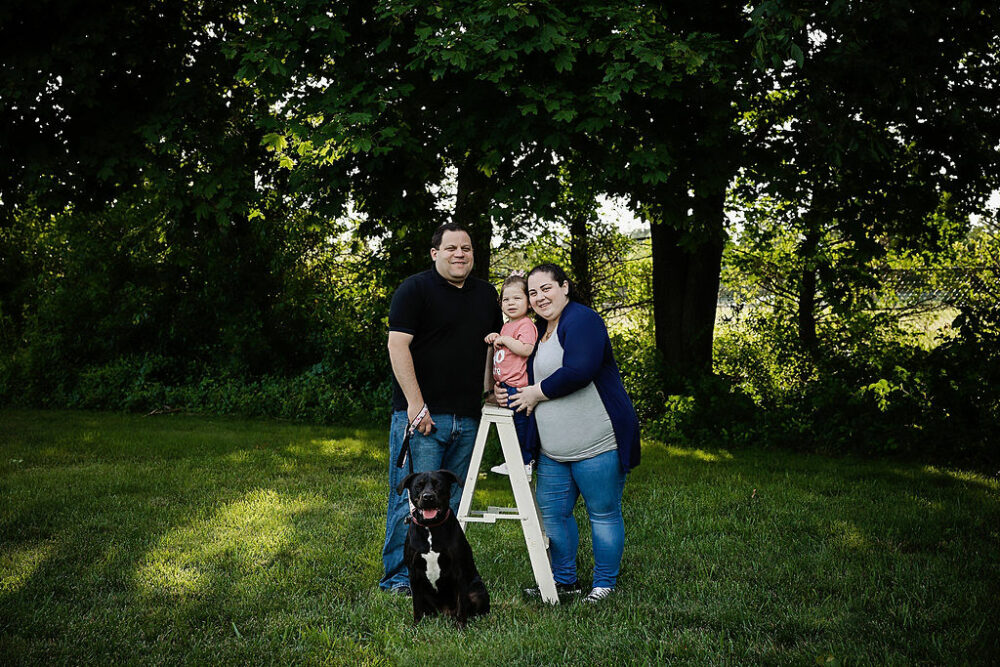 Family portrait of a father and mother with their daughter, and between them and family dog sitting for their professional baby photos taken for their daughters mile stone photography session in Morristown, New Jersey.