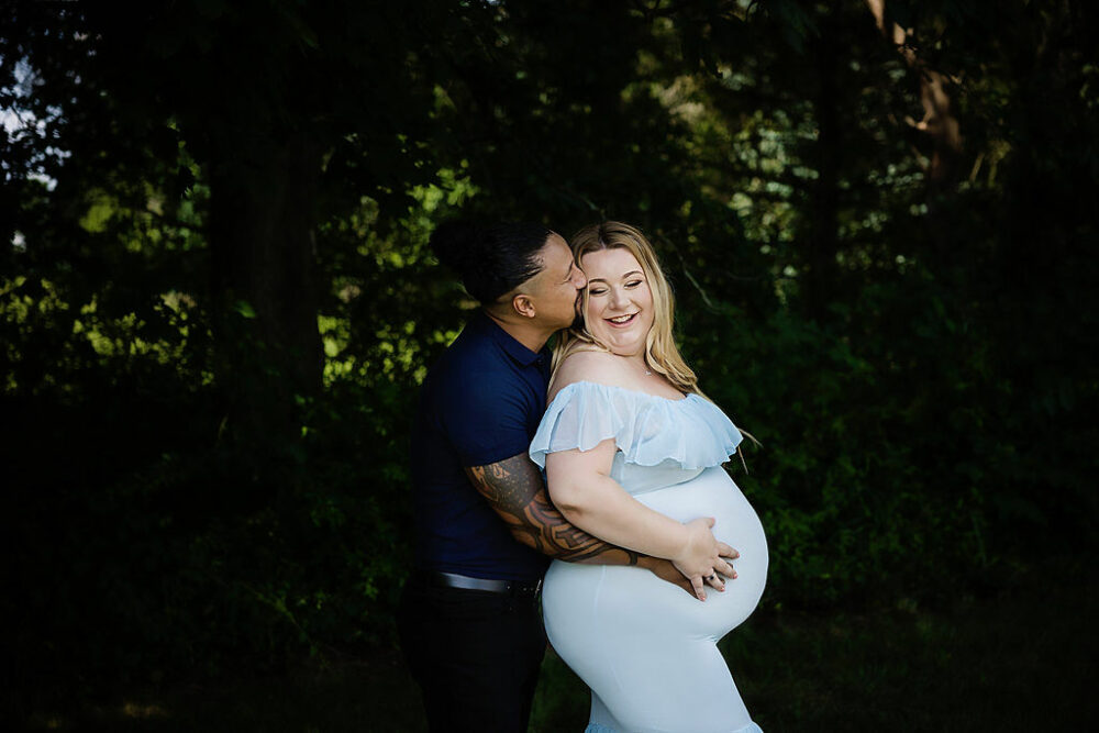 Husband kissing his pregnant wife on cheek while standing behind her in a prom pose for her Blue Maternity and Newborn Session in Camden, New Jersey.