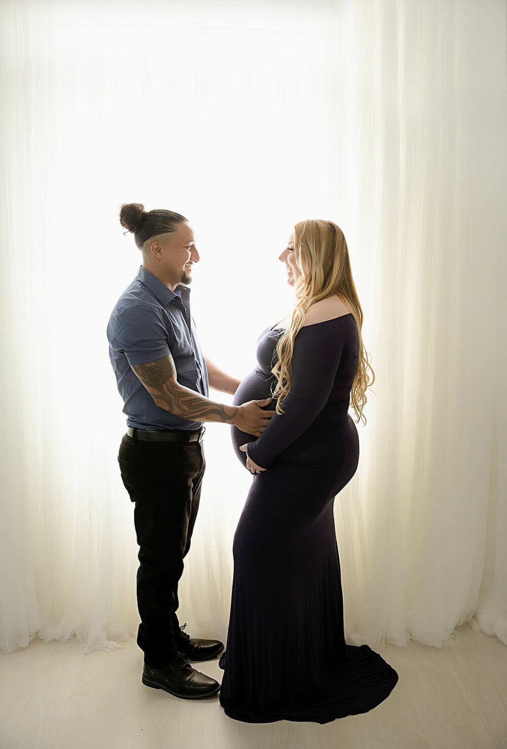 Husband and wife facing each other while holding pregnant belly against a backlit shot for their couple maternity photos taken in Moorestown, New Jersey.