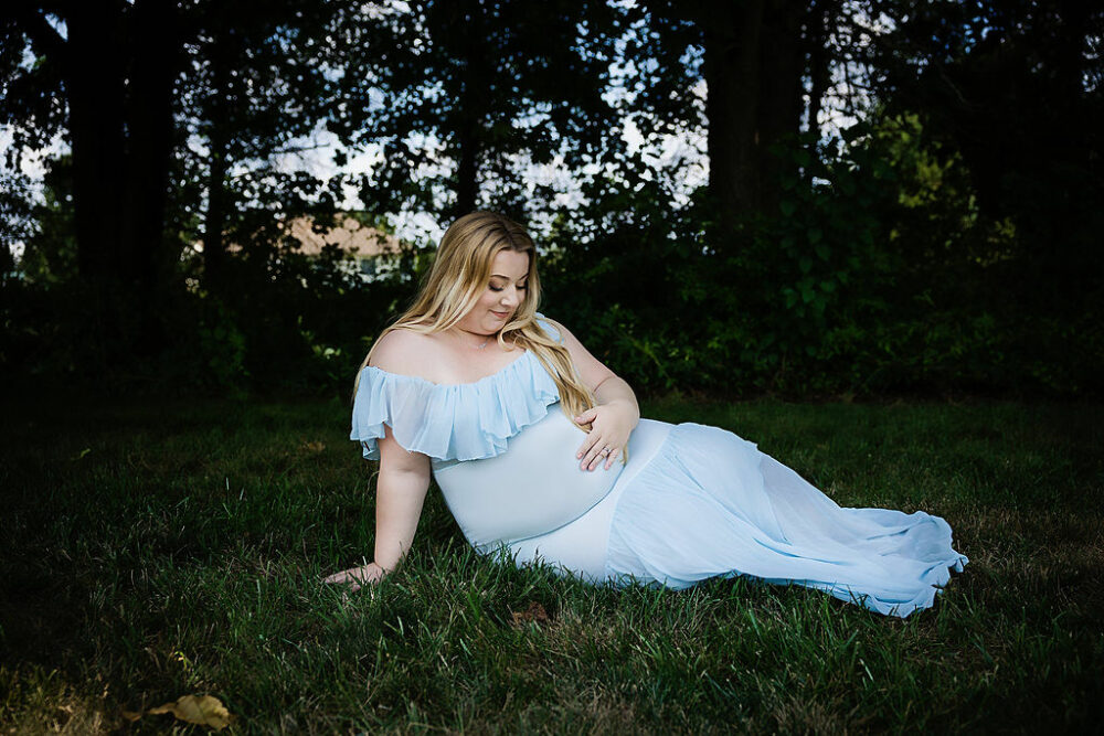 Pregnant woman sitting on grass, outdoors, holding and looking at her stomach for her blue maternity session in Pemberton, New Jersey.