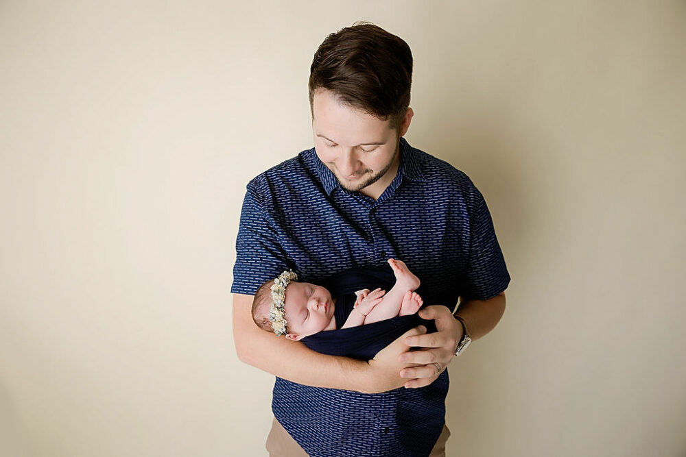 Man looking down and smiling at his newborn daughter, which he’s holding lovingly in his arms for her NICU in-studio newborn session in Pemberton, New Jersey.