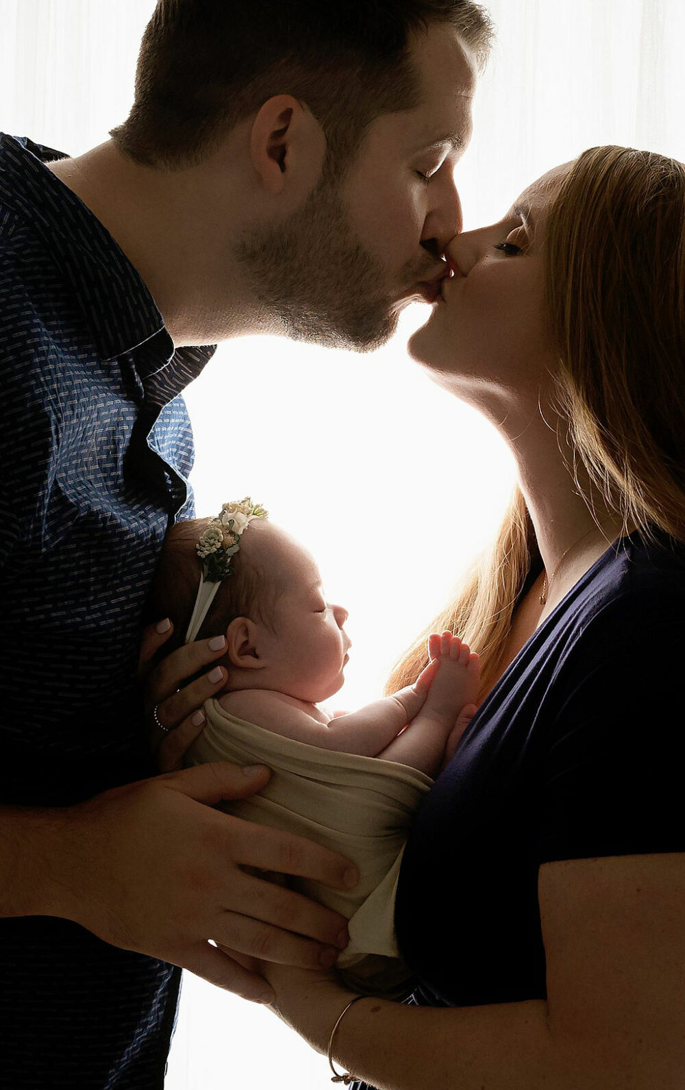 Close-up of man and woman kissing and holding newborn baby between them while she sleeps against black backdrop for newborn photography during their Nicu in-studio newborn session in Southampton, New Jersey.