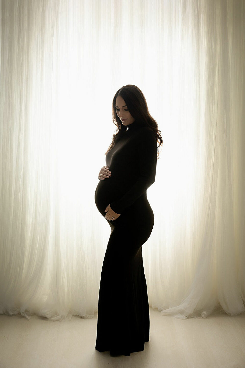 Pregnant woman wearing long dress and posing by holding her belly and looking down against light and bright backdrop for her black-and-white maternity session in Southampton, New Jersey.