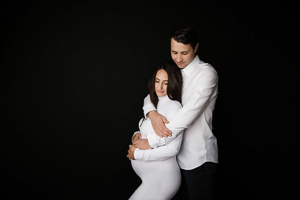 Man standing behind his pregnant wife wearing cute maternity photo shoot outfits for a black and white photography session in Monmouth County, New Jersey.