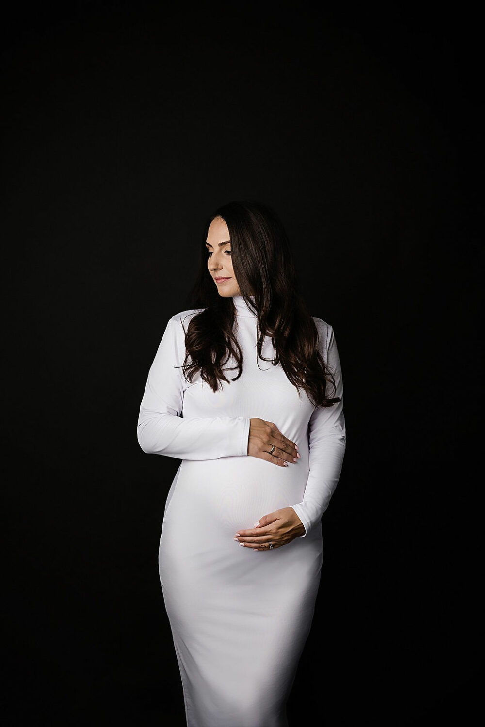 Woman holding her belly standing facing toward camera looking off to the side for her maternity photo shoot poses taken in studio for her black and white maternity session in Hamilton, New Jersey.