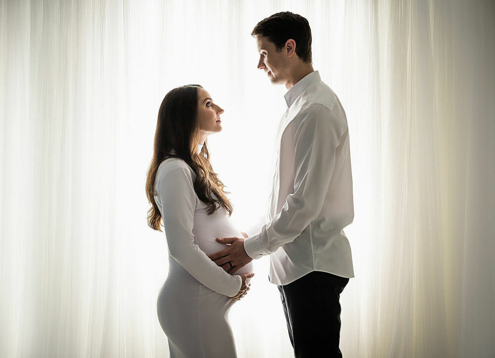 Men and women facing each other and looking at one another while holding women's belly for her white and light classic maternity session taken in Morristown, New Jersey.