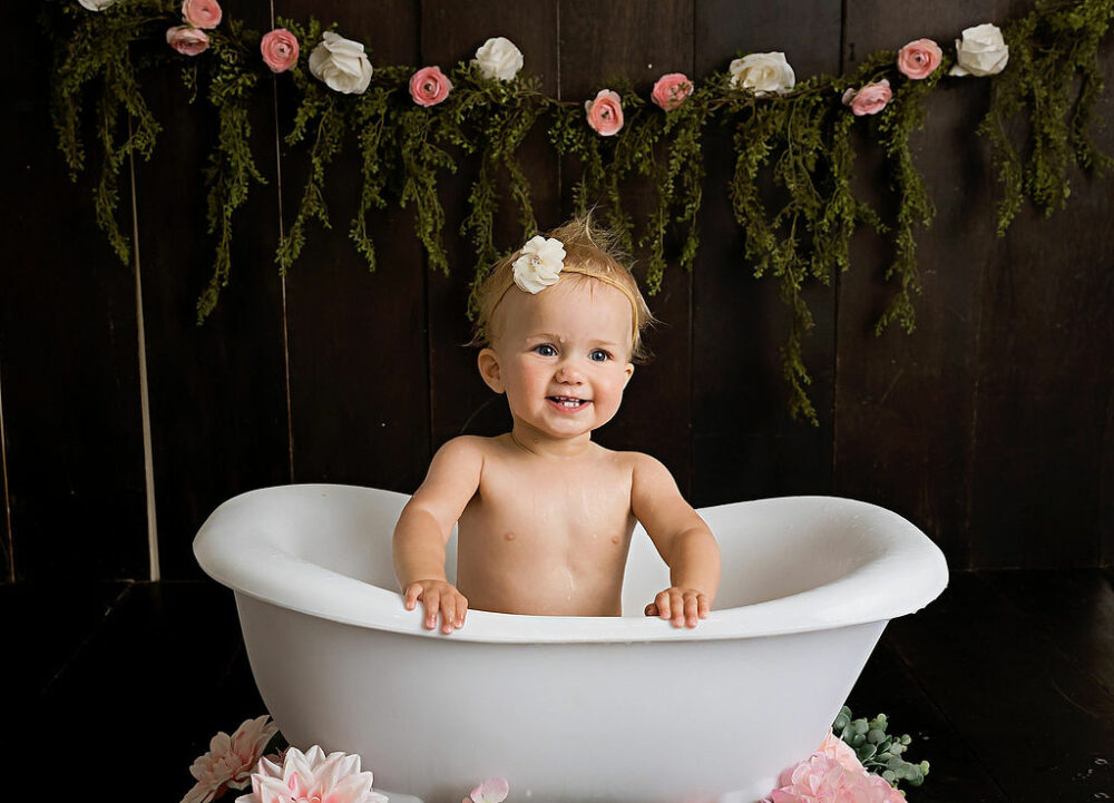 Toddler girl, smiling at camera and sitting tub photography prop adorned with flowers for her first birthday photography in Eastampton, New Jersey.