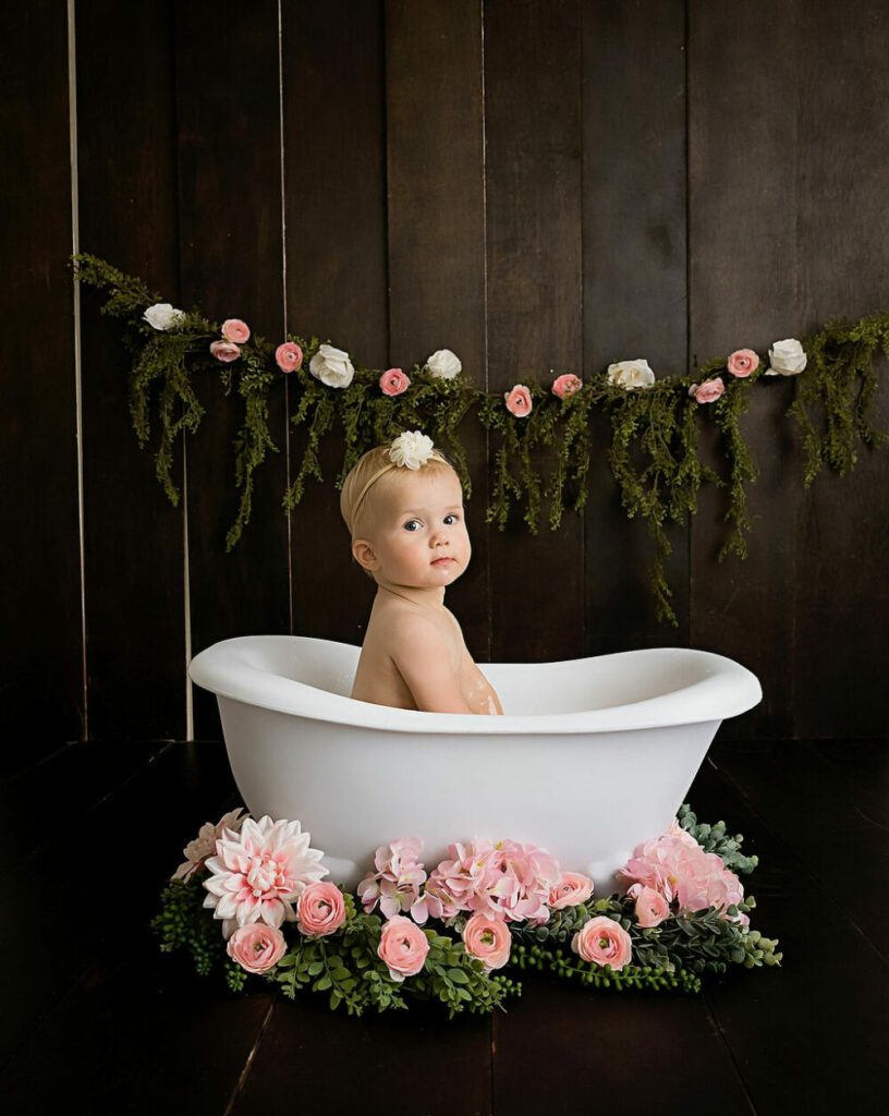 Cute baby portrait of a toddler girl sitting in photography prop bathtub, adorned with greenery and flowers for her in studio watermelon first birthday session in Monmouth County, New Jersey.