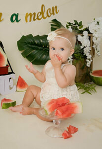 One year old girl, looking at camera and eating watermelon for first birthday ideas photo shoot taken in Mount Holly, New Jersey.
