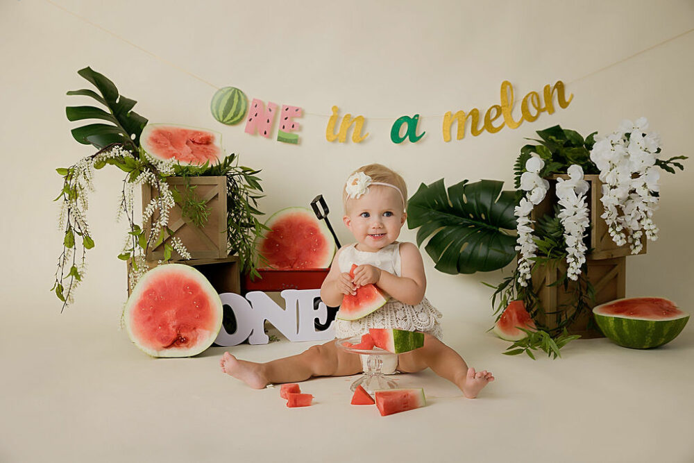 One year old girl, sitting and eating watermelon in professional photography studio for her watermelon First Birthday session taken for her baby milestones in Westhampton, New Jersey.