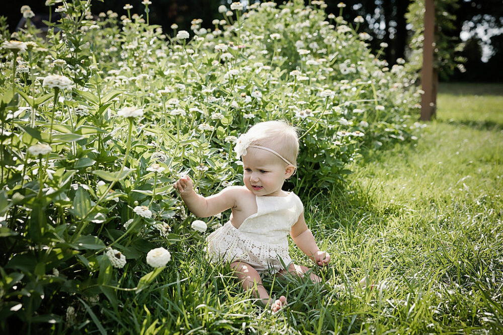 One year old girl, sitting in grass next to flower garden, wearing dress and headband for first birthday ideas photo shoot in Cherry Hill, New Jersey.