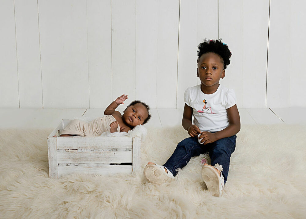 Toddler girl sitting on floor next to her baby sister and photography prop both looking at camera for her neutral in-studio newborn session in Mount Holly, New Jersey.