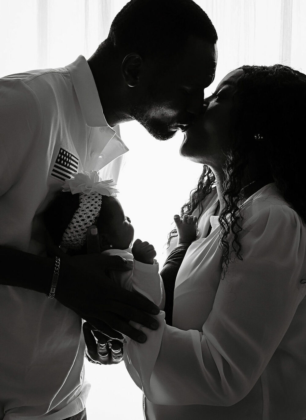 Man and woman kissing while holding their newborn baby girl in between them taking during their neutral in studio newborn session in Monmouth County, New Jersey.