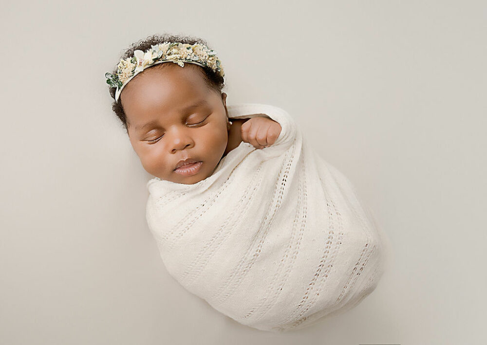 Close up of newborn girl wrapped in white blanket wearing matching headband while sleeping on beanbag photography prop for her baby picture ideas photo shoot taken in Cherry Hill, New Jersey.
