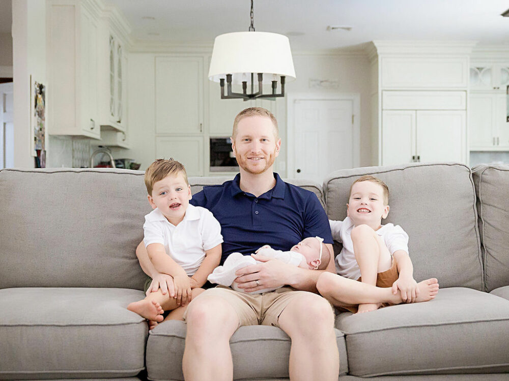 Man and his two sons sitting on their living room couch, while he is holding newborn girl, and his arms are smiling at the camera for their newborn family photos taken in Southampton, New Jersey.
