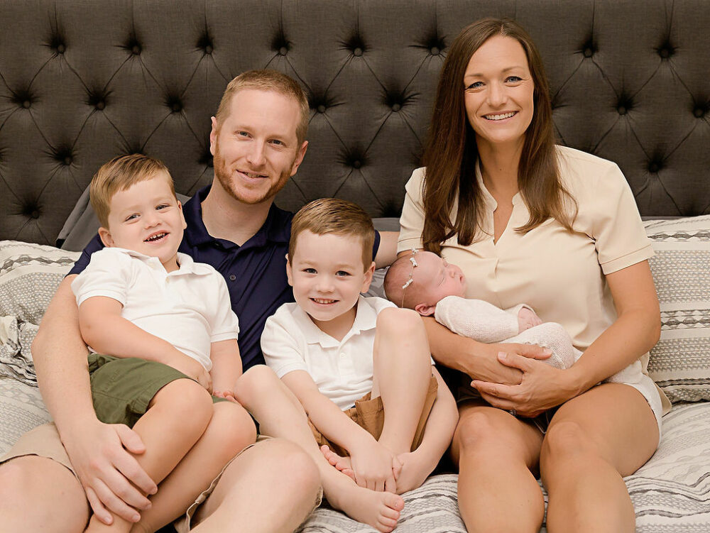 Close up of a man and a woman and their two toddler sons, and Sleepy newborn girl in master bedroom and smiling at camera for their in-home newborn session, taken in Westhampton, New Jersey.