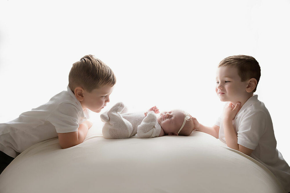 Two toddler boys facing each other, while looking at their newborn baby sister, on beanbag photography prop for her and her newborn session, taken in Camden, New Jersey.