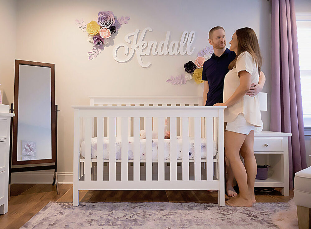 A man and women embracing each other, standing next to newborn daughters crib in her nursery with purple accents for their purple and home newborn session taken in Vincentown, New Jersey.