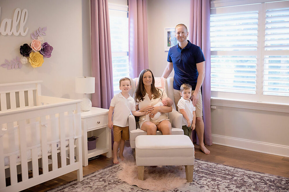 Woman sitting on nursing chair, holding her newborn girl while her husband and two kids are standing around her posing for family portraits and baby nursery for their purple in Home newborn session taken in Cherry Hill, New Jersey.