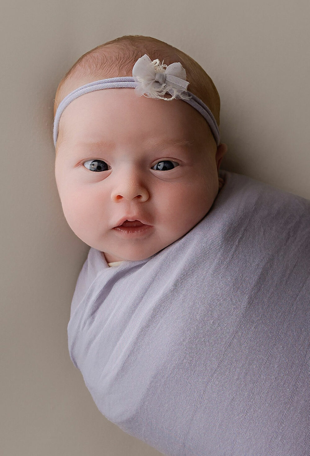 Close-up, infant portrait of newborn girl, wrapped and wearing headband, looking at camera for her professional baby pics taken during her purple in Home newborn session in Mount Laurel, New Jersey.