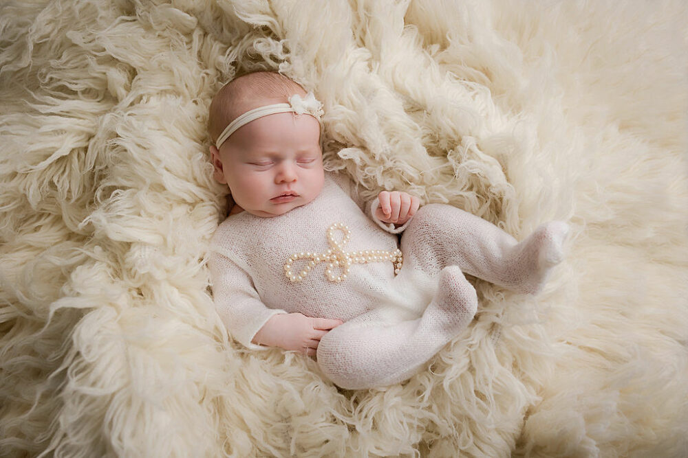 Infant girl sleeping on textured blanket, wearing headband pearls and knit outfit for her newborn pictures taken in Mount Holly, New Jersey.