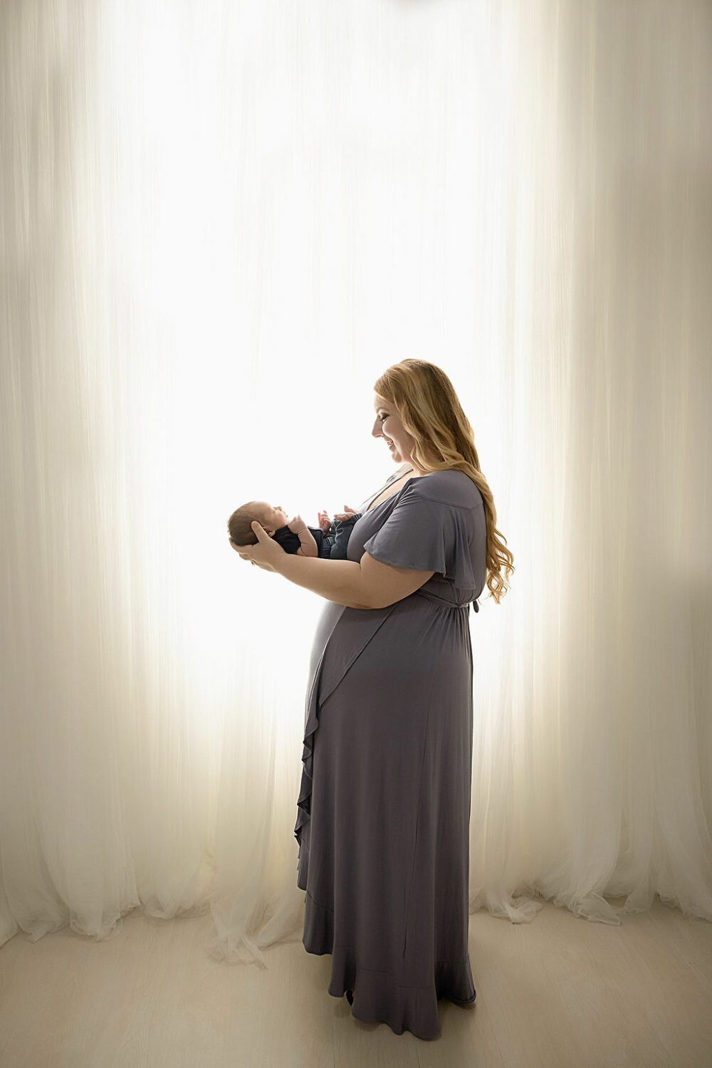 Side view of a woman standing and holding her newborn son in her arms against a backlit background for their blue maternity session taken in a professional studio in Southampton, New Jersey.