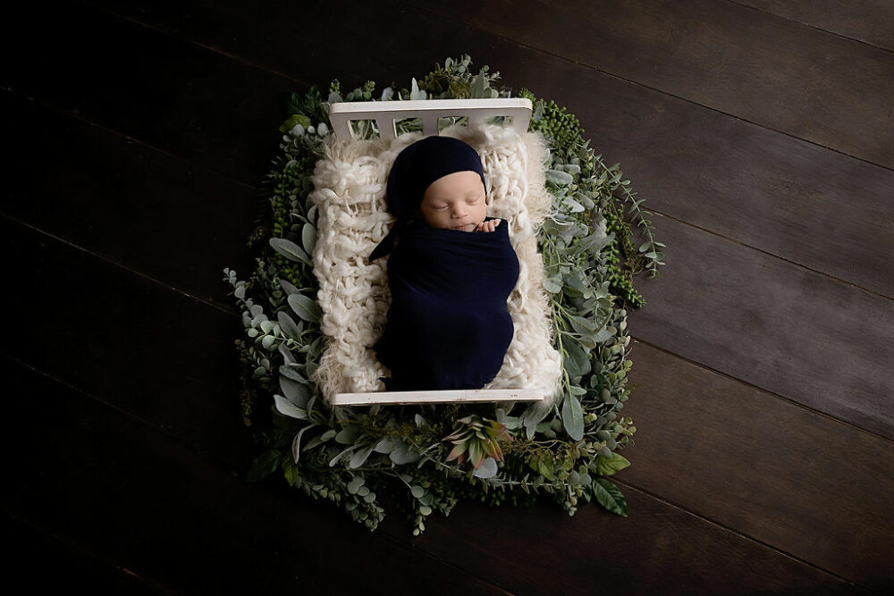 Newborn boy sleeping on crib photography prop wrapped in swaddling on textured blankets and greenery for his professional blue newborn session taken in Morristown, New Jersey.