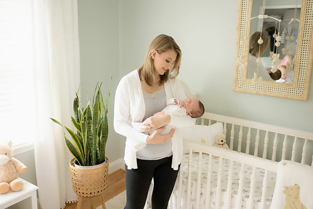 Woman holding her infant in her arms, while looking down at him, while he is sleeping, standing in front of his crib in his nursery, for a bright in Home Newborn session, taken in Hamilton, New Jersey.