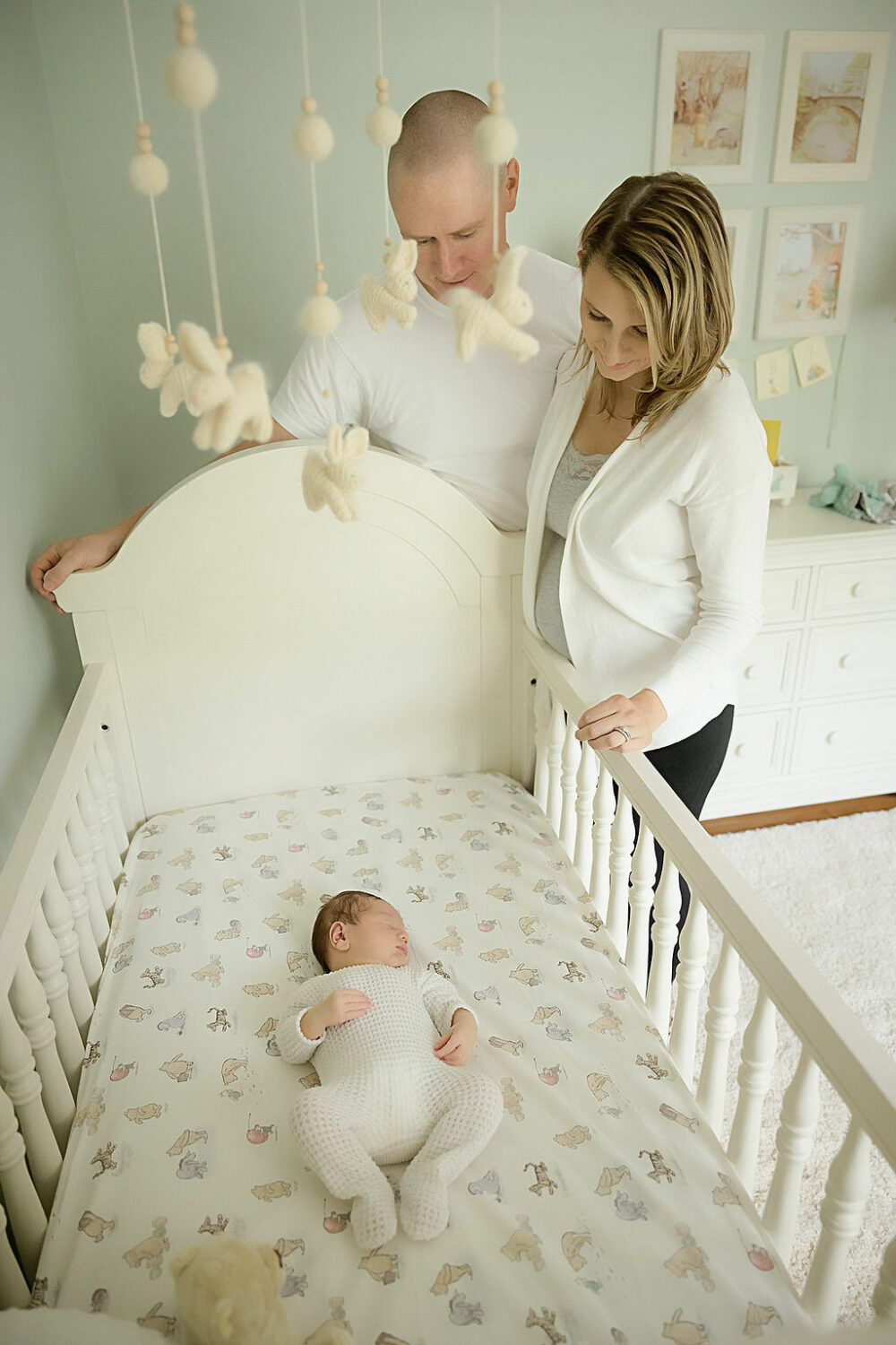New parents looking down at Sleepy infant boy in his crib posing in nursery for their bright, in-home newborn session, taken in Monmouth county, New Jersey.