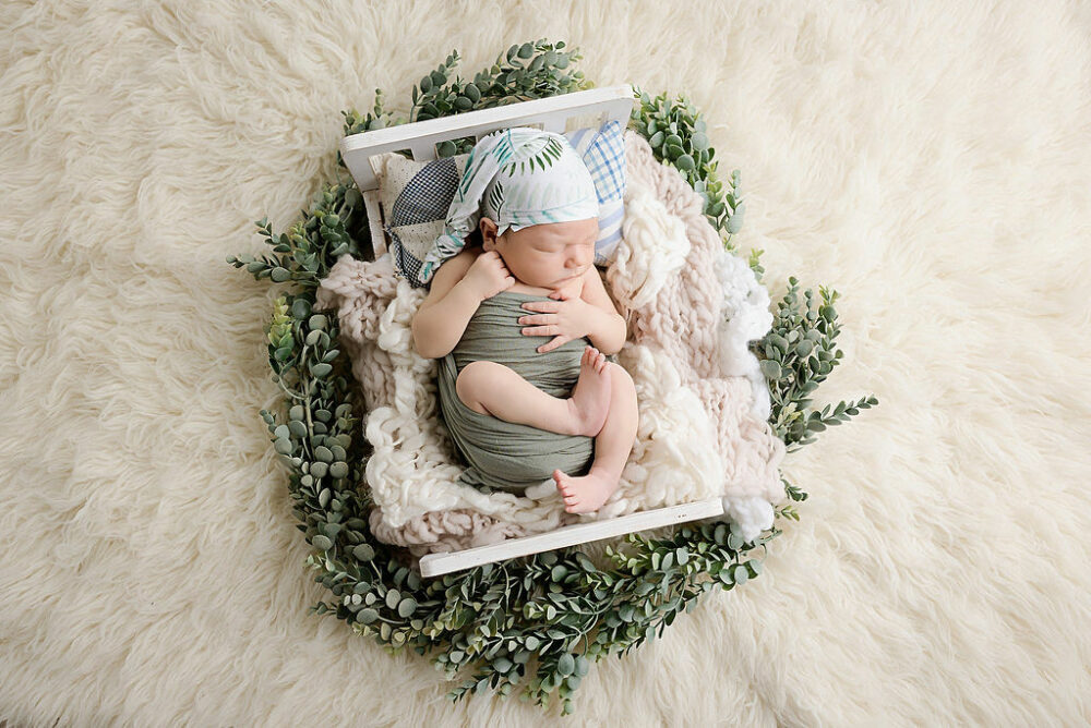 Sleepy newborn boy on swaddled and sleeping on newborn photography prop adorned with greenery and blankets for his bright in-home Newborn session taken in New Egypt, New Jersey.