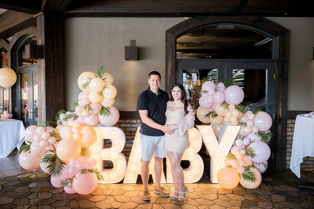 Man and woman standing next to each other smiling a camera while holding her belly standing in front of large sign and restaurant and you for their baby shower event in Monmouth County, New Jersey.