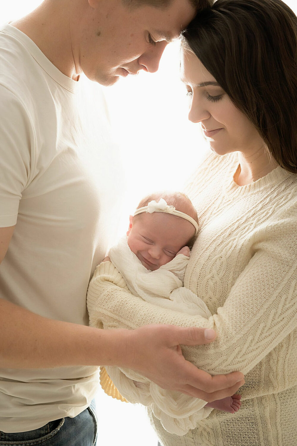Close-up of a man and a woman touching foreheads, while looking down at smiling newborn daughter in between them for dare in studio, newborn photo, shoot in Burlington, New Jersey.