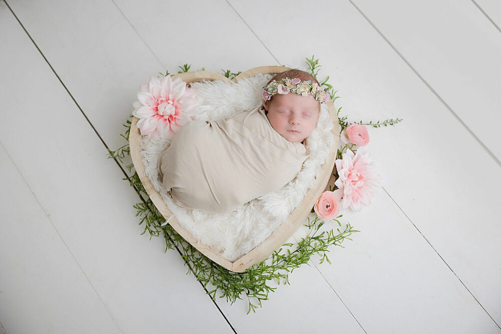 Newborn girl, sleeping and swaddled, wearing headband on newborn photography prop with flowers taken during her pink newborn session in Camden, New Jersey.
