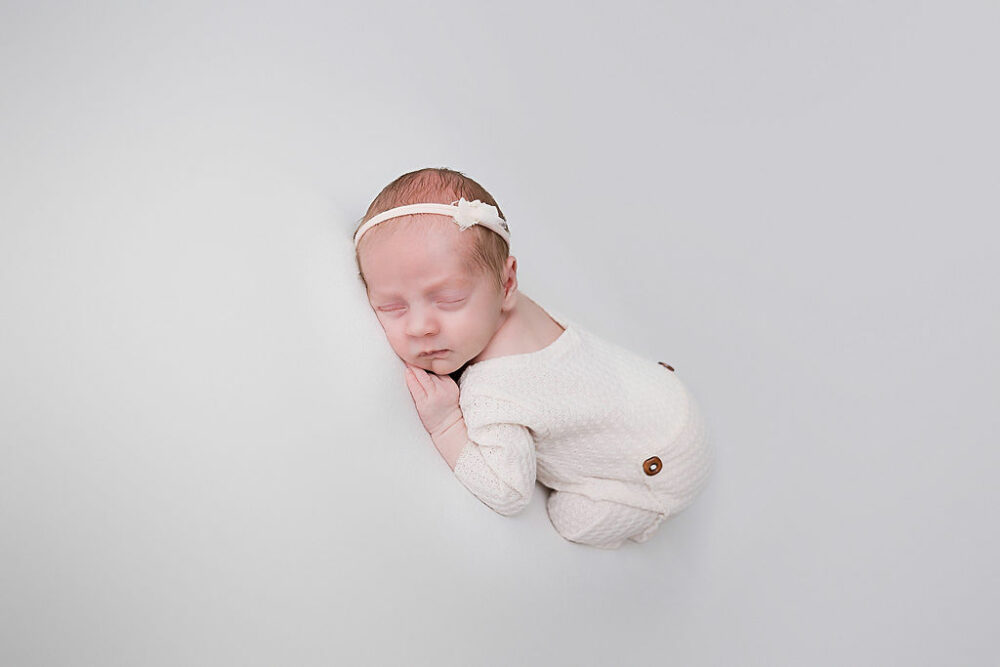 Newborn girl, sleeping on her tummy, wearing cute outfit and headband against white backdrop for her baby pictures taken in Westhampton, New Jersey.