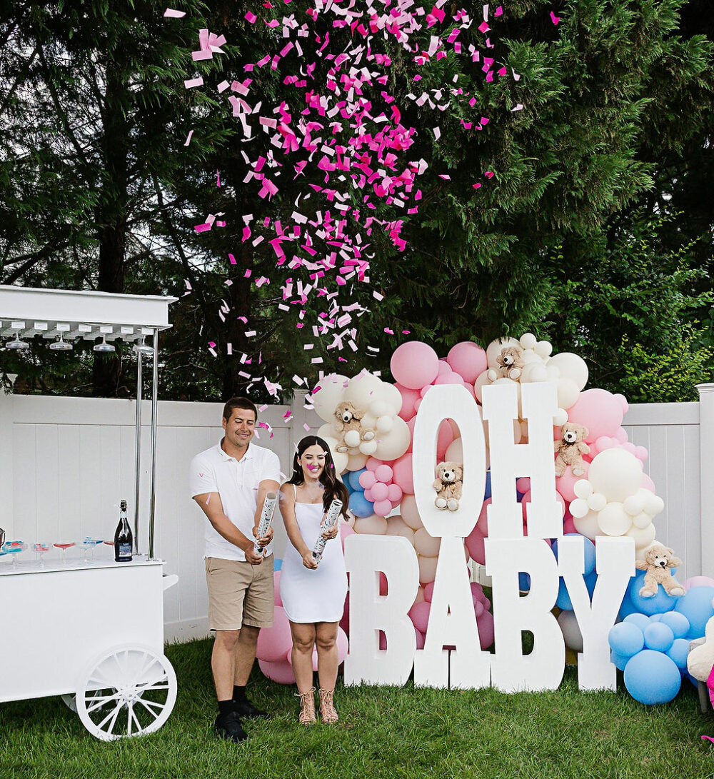 Man and woman standing next to large gender, reveal sign with balloons and a cart, smiling at camera for their gender reveal photo, shoot, and Morristown, New Jersey.