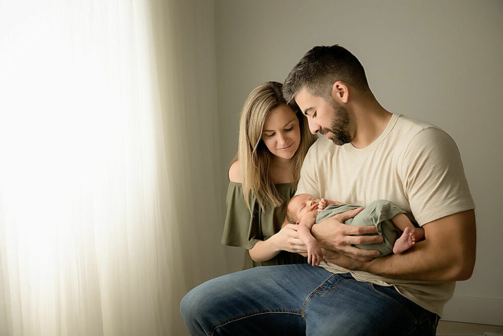 Man sitting down and holding his infant son while his wife is kneeling next to him looking at him as well for their green in studio newborn session taken in Pemberton, New Jersey.