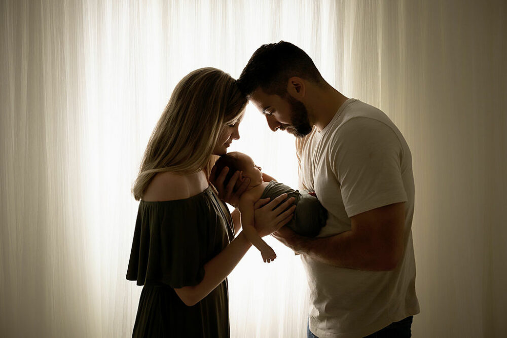 Woman and man facing each other while holding newborn boy in between them and touching foreheads taken for their family portrait ideas taken in studio in Morristown, New Jersey.