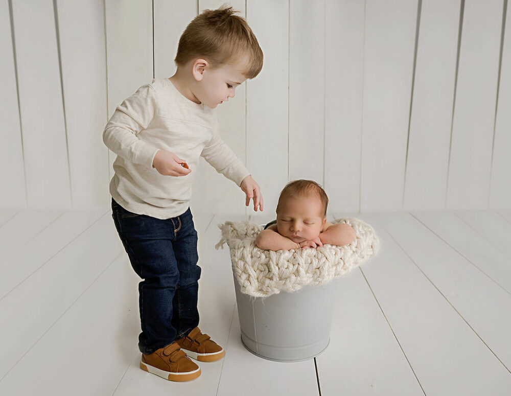 Toddler boy standing next to sleepy infant brother while he is in a photography prop bucket with blanket for their green in studio newborn session in Hamilton, New Jersey.