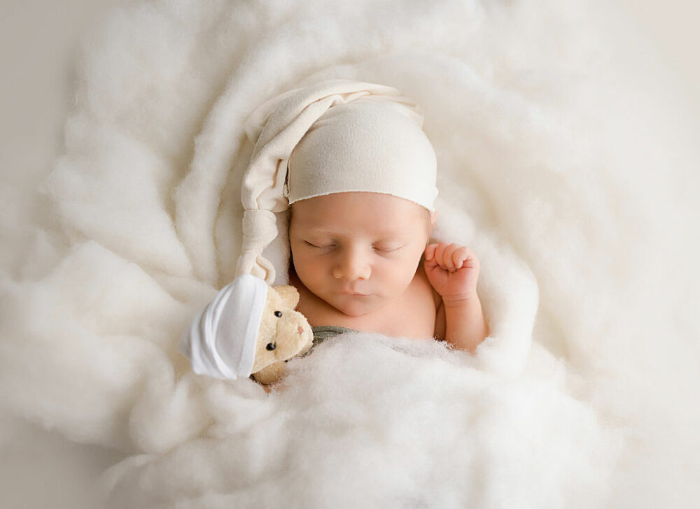 Newborn boy sleeping next to his plushy surrounded by blanket photography prop with one hand by his head for his professional baby pics taken in studio in Mount holly, New Jersey.