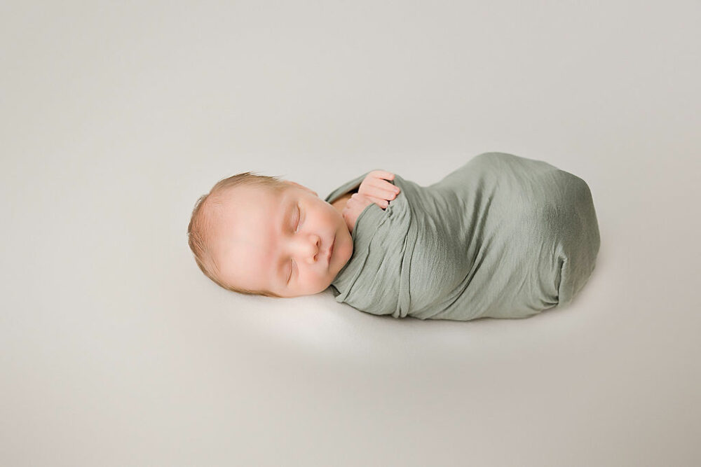 Infant boy sleeping on beanbag photography prop swaddled with hands showing for his green in-studio newborn session in Camden, New Jersey.