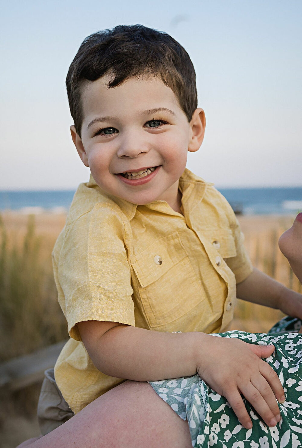 Toddler boy smiling and looking at camera as he's being held by his mother during their lifestyle family big session in Ocean City, New Jersey.